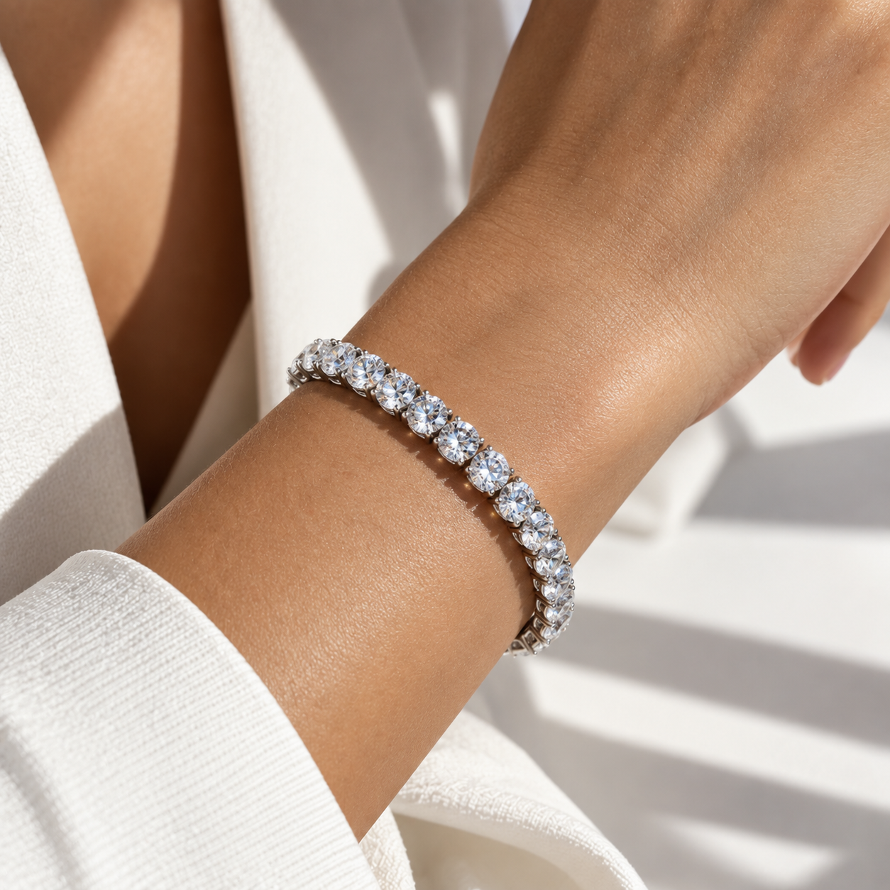 A close-up of a woman's wrist wearing a sparkling diamond tennis bracelet in a silver-toned setting, styled with a white textured top under bright natural light.
